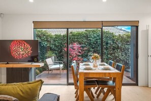 Ein Essbereich mit einem Tisch für vier Personen, einem Fernseher auf einem Schrank und Blick auf einen Garten durch große Glasüren.