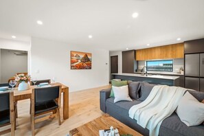Ein modernes Wohnzimmer mit grauem Sofa, einem Holzesstisch und einer Küchenzeile mit Einbauofen und Kühlschrank.