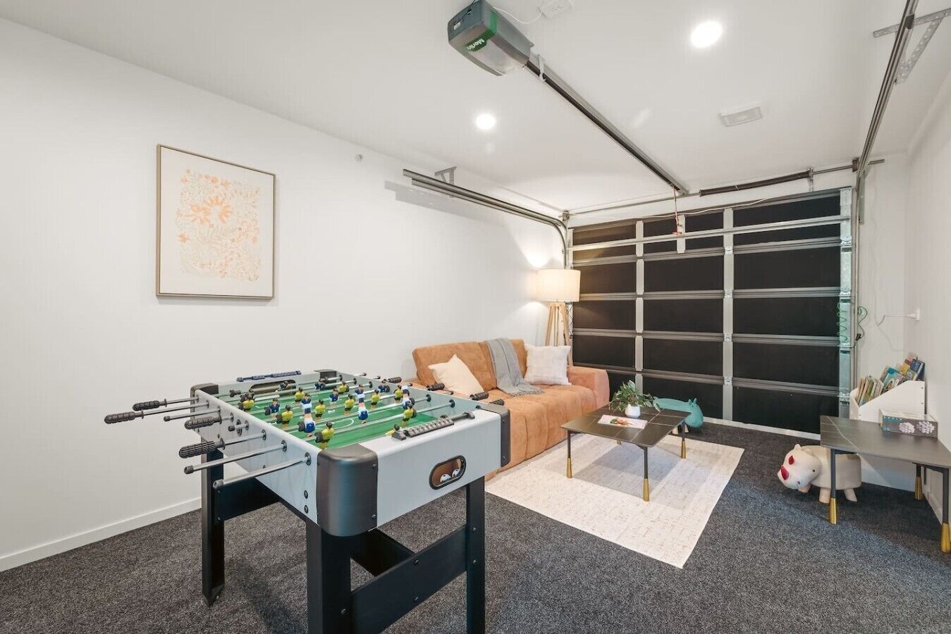 Une pièce avec un baby-foot, un canapé, une lampe, une petite table avec une plante et une fenêtre avec des stores.