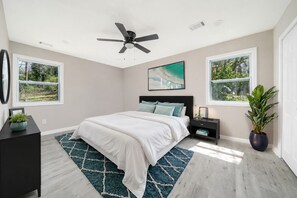 Une chambre à coucher comprenant un lit, un ventilateur de plafond, deux fenêtres, une table de chevet, une commode et une plante en pot.