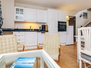A compact kitchen with white cabinets, a coffee machine, and a refrigerator. There are two chairs with cushions and a dining table.