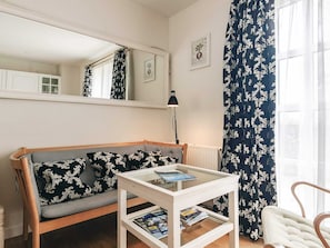 A room with a sofa, a glass-top table with magazines, a lamp, a mirror, and a window with curtains.