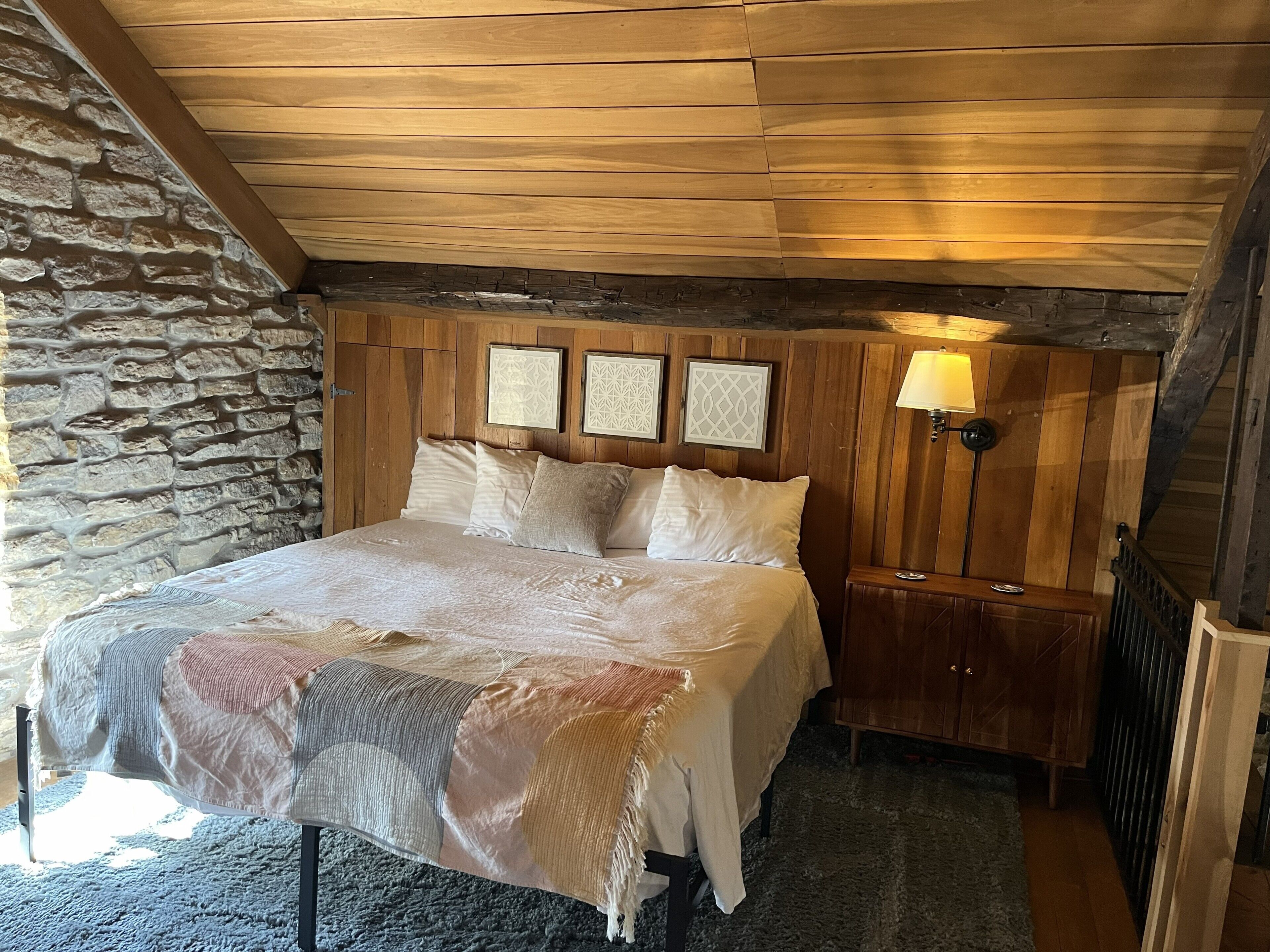 A bedroom with a stone wall, wooden ceiling, a bed with white bedding, and a nightstand with a lamp.