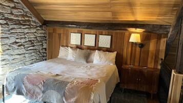 A bedroom with a stone wall, wooden ceiling, a bed with white bedding, and a nightstand with a lamp.