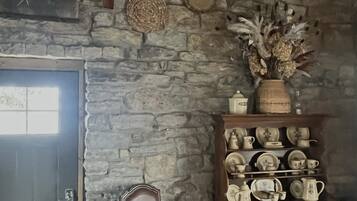 Ett rustikt rum med en vägg av sten, en trästol med en kudde, ett skåp med keramikföremål och en tavla på väggen.