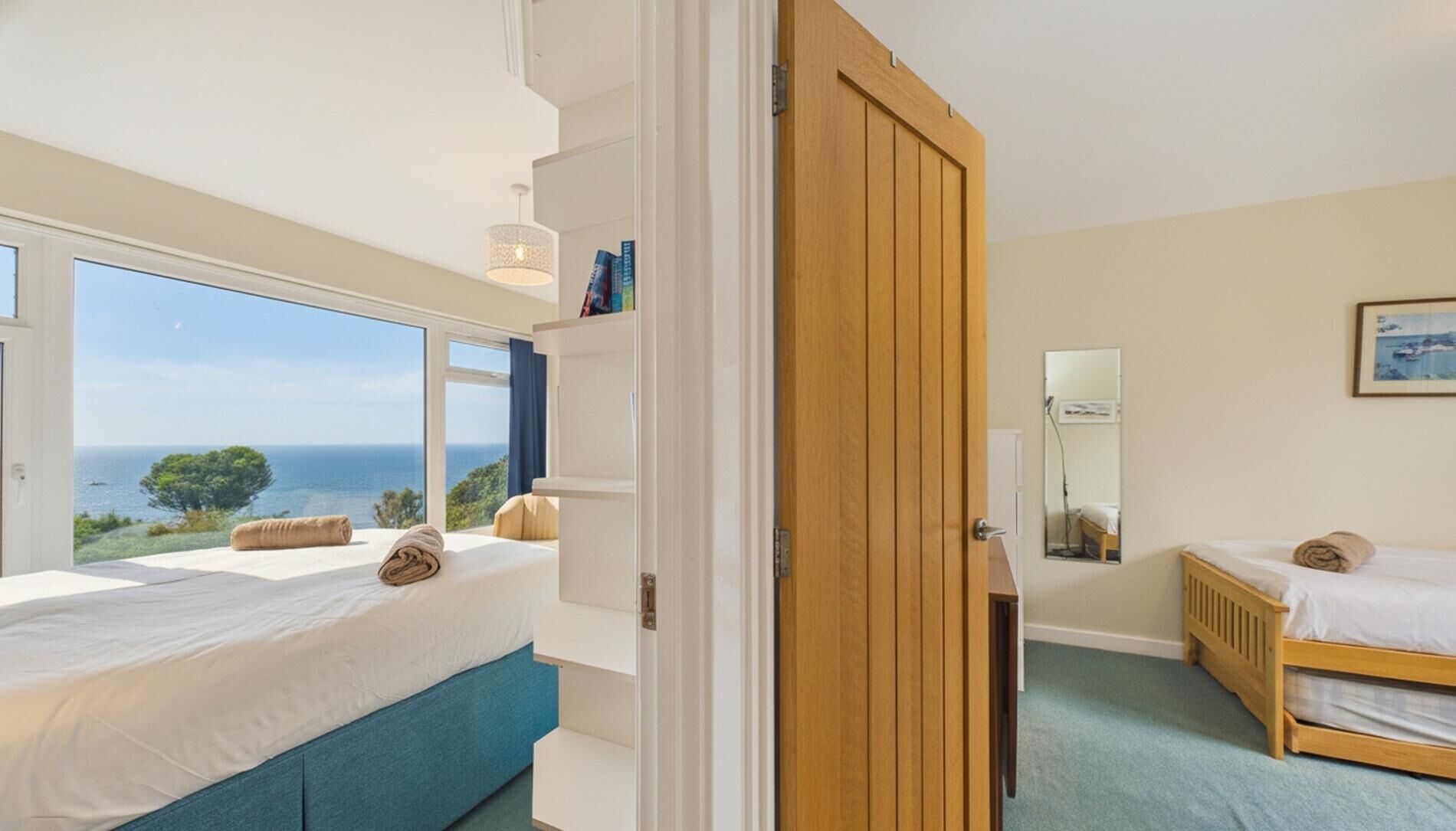 A bedroom with two beds, a large window with a sea view, a wooden door, and a mirror on the wall.