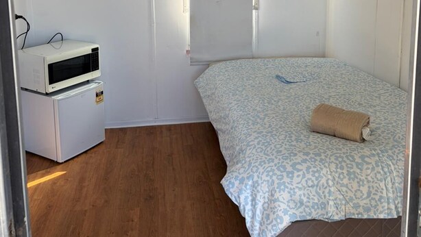 Een kleine eenpersoonskamer met een houten vloer, een bed met een bedekking met patroon, een witte verwarming en een airconditioning.