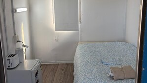 Een kleine, compacte kamer met een bed, nachtkastje met lamp, stopcontact en raam.