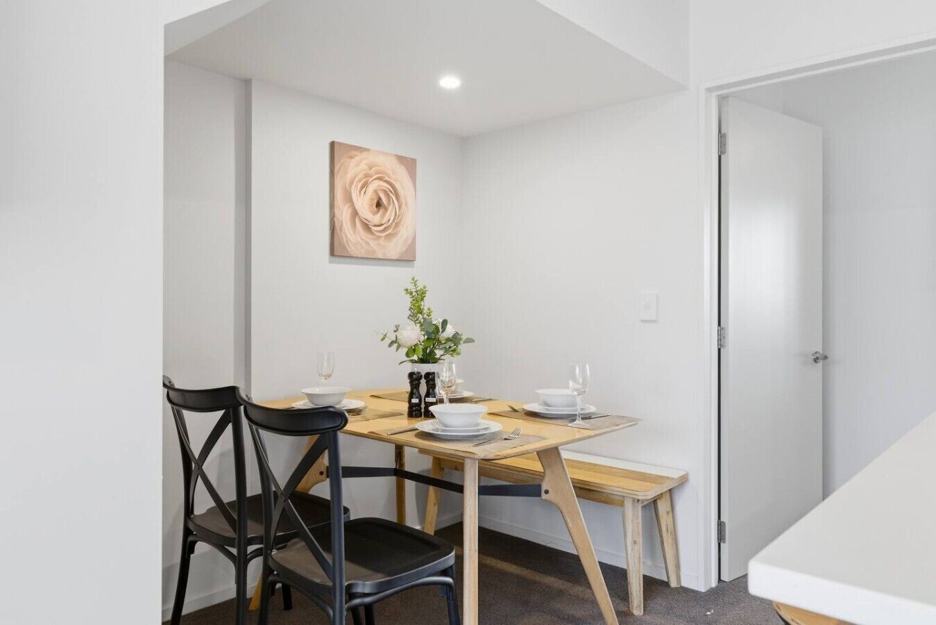 Uma sala de jantar com uma mesa de madeira, duas cadeiras pretas, um vaso com flores e um quadro na parede.