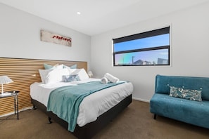 Ein Schlafzimmer mit einem Bett, einem blauen Sofa, einem Nachttisch, einer Lampe, einem Fenster mit Stadtblick und einem Wandbild.