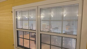 Ein Fenster mit Jalousien, eine gelbe Wand und eine sichtbare Backsteinmauer draußen.