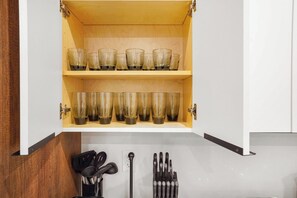 A kitchen cabinet with two shelves holding clear glasses, a utensil holder with black utensils, and a metal utensil holder with metal utensils.