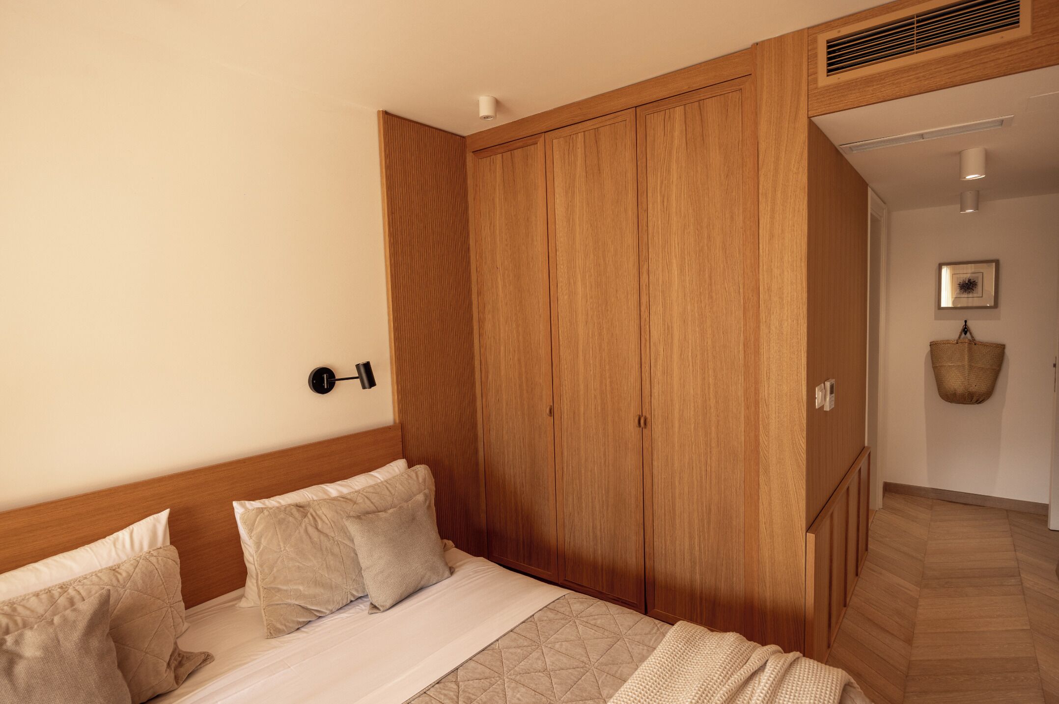 Un dormitorio con un armario de madera, una cama con ropa de cama color beige y una lámpara fijada a la pared.