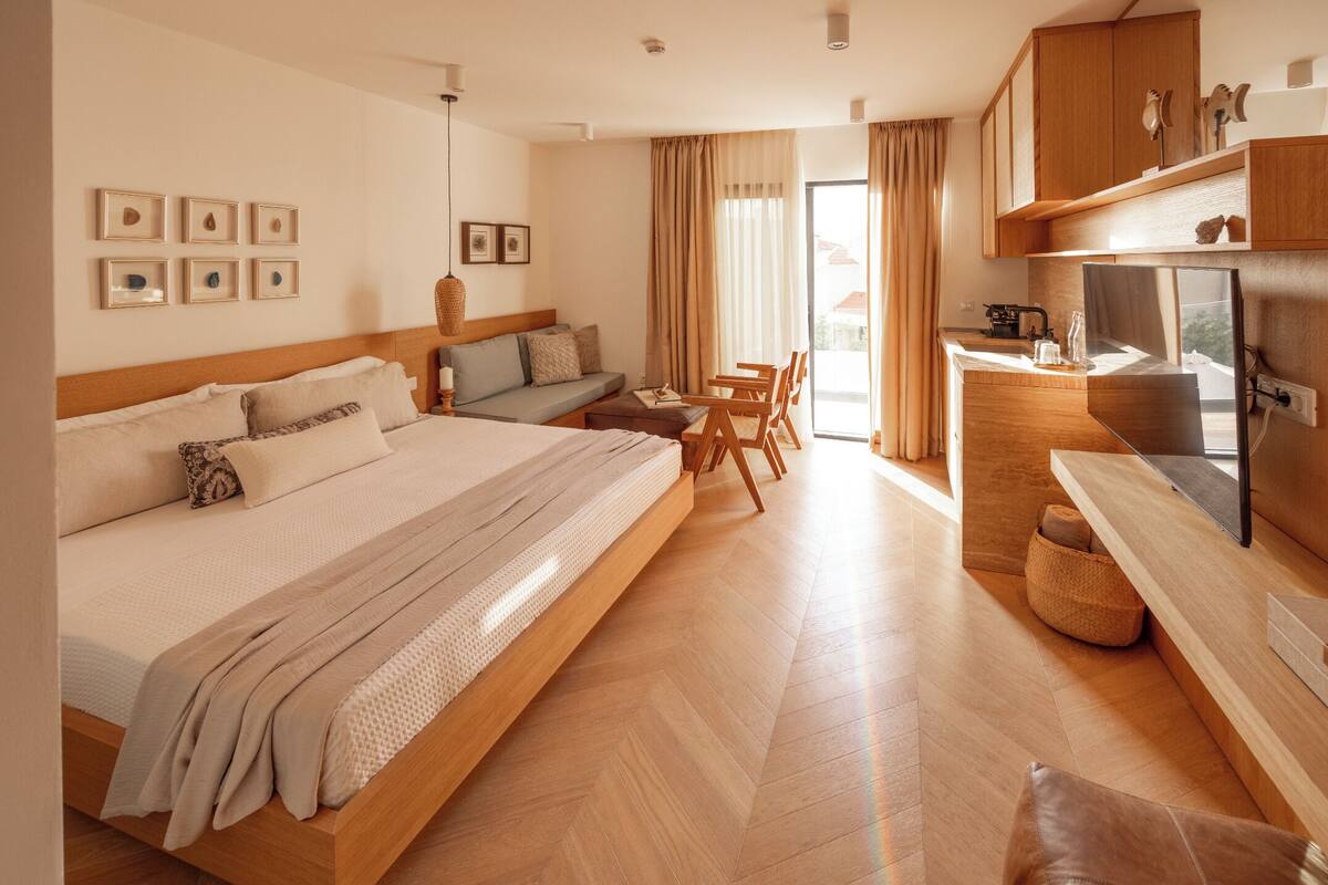 A modern hotel room with a large bed, a sofa, a dining area, and a kitchenette.