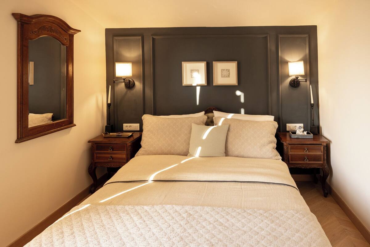 A neatly made bed with a quilt, two bedside tables with lamps, a mirror, and two framed pictures on the wall.