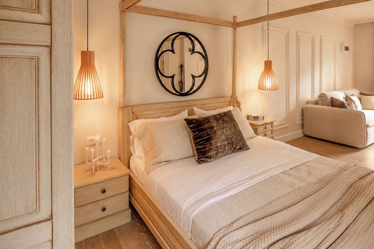 A bedroom with a wooden bed, bedside tables, a wall-mounted light fixture, and a sofa in the adjacent room.