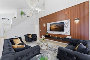 A modern living room with a large TV, dark sofas, a glass coffee table, and a wooden accent wall. - 9 Minutes to Disney Pool + Theater + Game Room (Kissimmee)