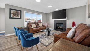 A living room with a brown leather sofa, blue chairs, a glass coffee table, a fireplace, and a TV mounted on the wall.