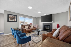 A living room with a brown leather sofa, blue chairs, a glass coffee table, a fireplace, and a TV mounted on the wall. - Luxury Urban Oasis in Prime Location (Airdrie)
