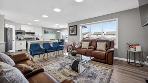 A modern living room with a brown leather sofa, blue chairs, a glass coffee table, and a kitchen in the background.