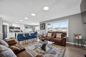 A modern living room with a brown leather sofa, blue chairs, a glass coffee table, and a kitchen in the background. - Luxury Urban Oasis in Prime Location (Airdrie)