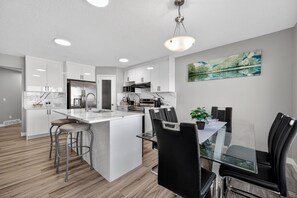 A modern kitchen with white cabinets, a central island, and a dining area with black chairs. - Luxury Urban Oasis in Prime Location (Airdrie)