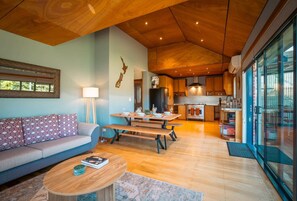 Ein modernes Wohnzimmer mit Sofa, Couchtisch und Essbereich, der in eine Küche mit Holzausbau und einem großen Fenster übergeht.