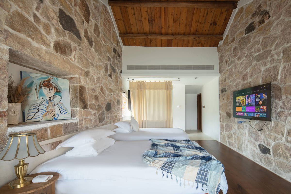 A bedroom with a stone wall, wooden ceiling, a bed with white bedding, a painting, a lamp, and a TV on the wall.