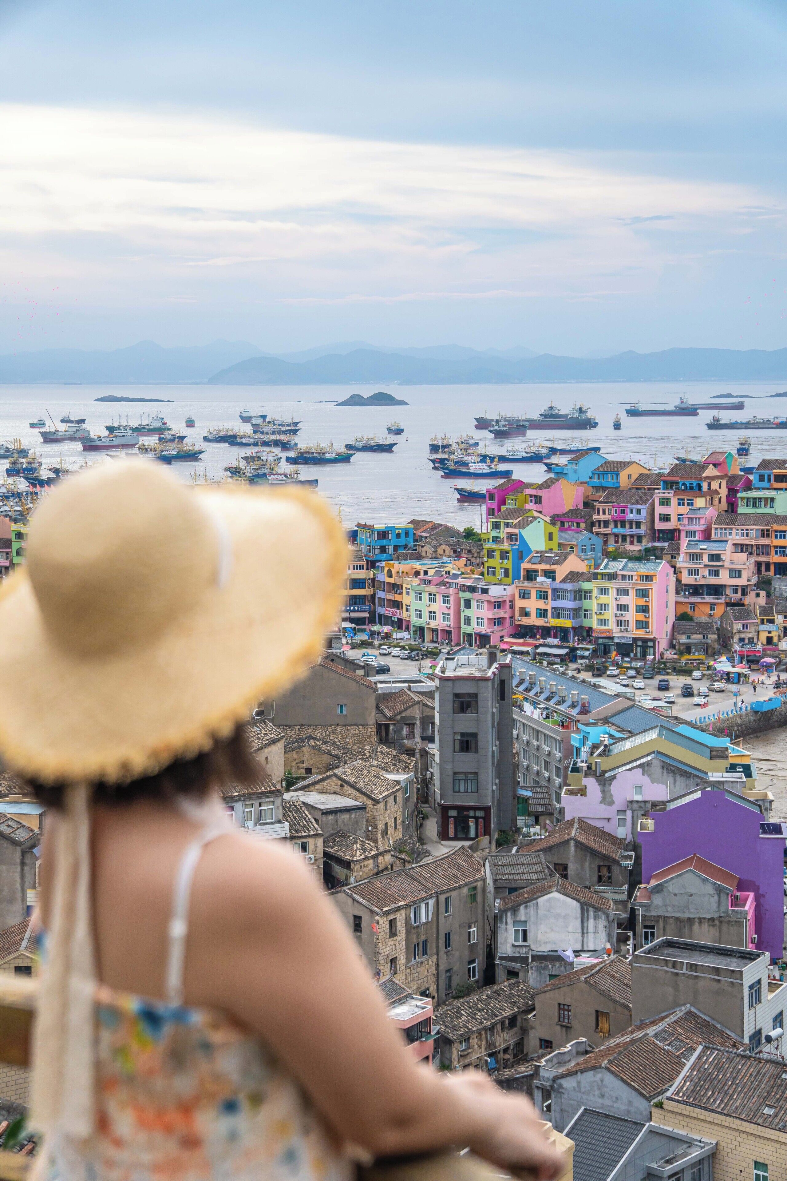 A woman in a hat overlooking a colorful coastal town with boats in the water.