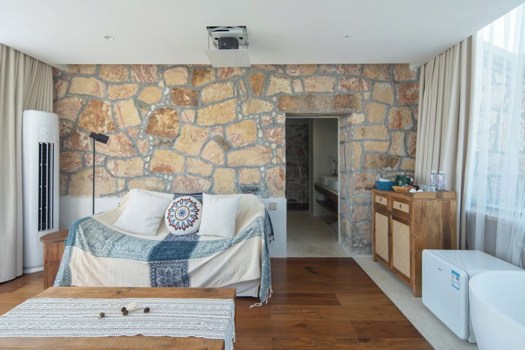 Un dormitorio con pared de piedra, cama con ropa de cama estampada, mesita de noche y vista a otra habitación.