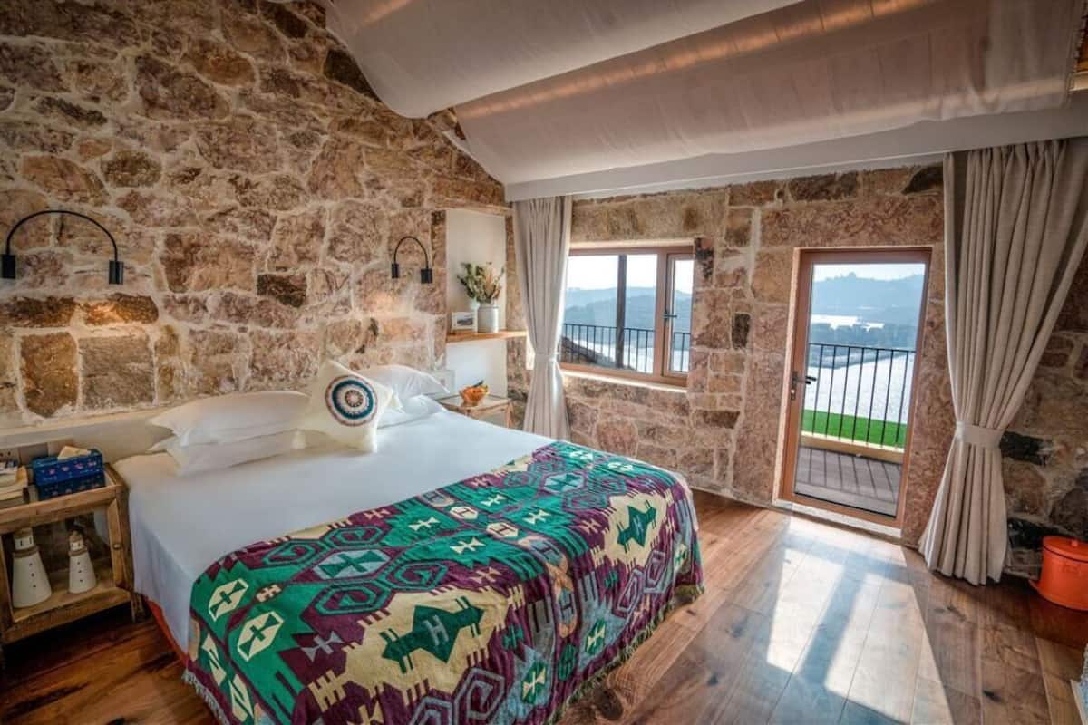A bedroom with a stone wall, a bed with a colorful blanket, a nightstand, a window with a view, and a balcony door.