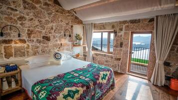Un dormitorio con pared de piedra, una cama con manta colorida, una mesita de noche, una ventana con vistas y una puerta al balcón.