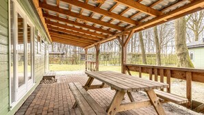Un espace pique-nique abrité, avec un banc et une table en bois, un sol en briques et une vue sur des arbres et un bâtiment en arrière-plan.