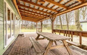Ein überdachter Picknickbereich mit einer Holzbänke und einem Tisch, Backsteinböden und Blick auf Bäume sowie ein Gebäude im Hintergrund.