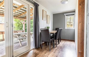 Ein Zimmer mit Holztisch und -stühlen, einem Fenster mit Blick auf Grünflächen und einer Tür, die zu einem Außenbereich mit einer Holzterrasse führt.