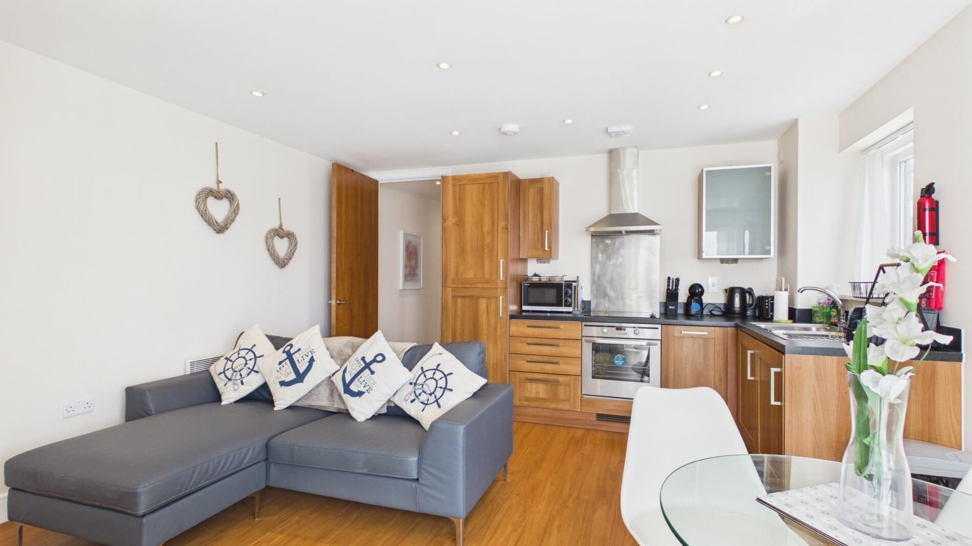 Een moderne woonkamer met een grijze hoekbank, houten kasten, een keuken met een magnetron, oven en een glazen tafel met een vaas.