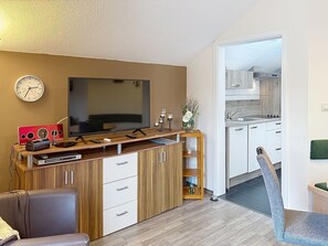 Una sala de estar moderna con una televisión de pantalla plana, un mueble bajo para equipo audiovisual y un comedor con una cocina al fondo.