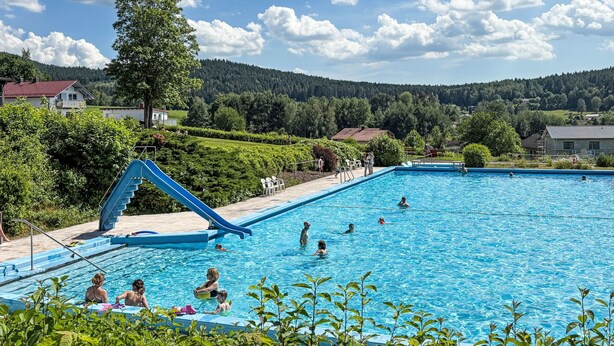 Una piscina con tobogán, rodeada de vegetación y casas, bajo un cielo despejado.