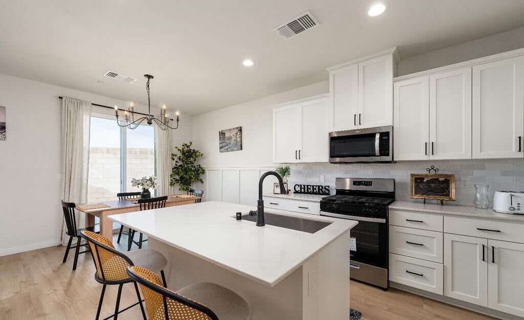 A modern kitchen with white cabinets, a central island with a sink, a microwave, and a stove. There is a dining area with chairs and a table.