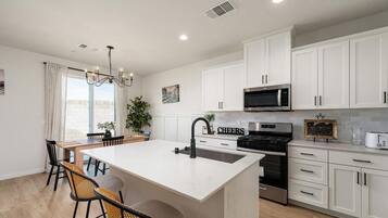 A modern kitchen with white cabinets, a central island with a sink, a microwave, and a stove. There is a dining area with chairs and a table.