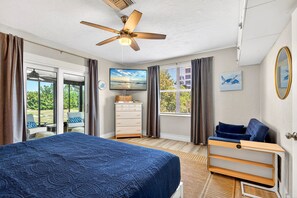 A bedroom with a bed, a ceiling fan, a dresser, a window with curtains, and a view of the outdoors.
