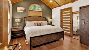 Een slaapkamer met een houten plafond, een bed met hoofdeinde, twee nachtkastjes met lampen, een raam met jaloezieën en een deur met spiegel.