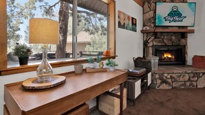 Una zona de estar acogedora con chimenea de piedra, una mesa de madera con lámpara y vistas a árboles.