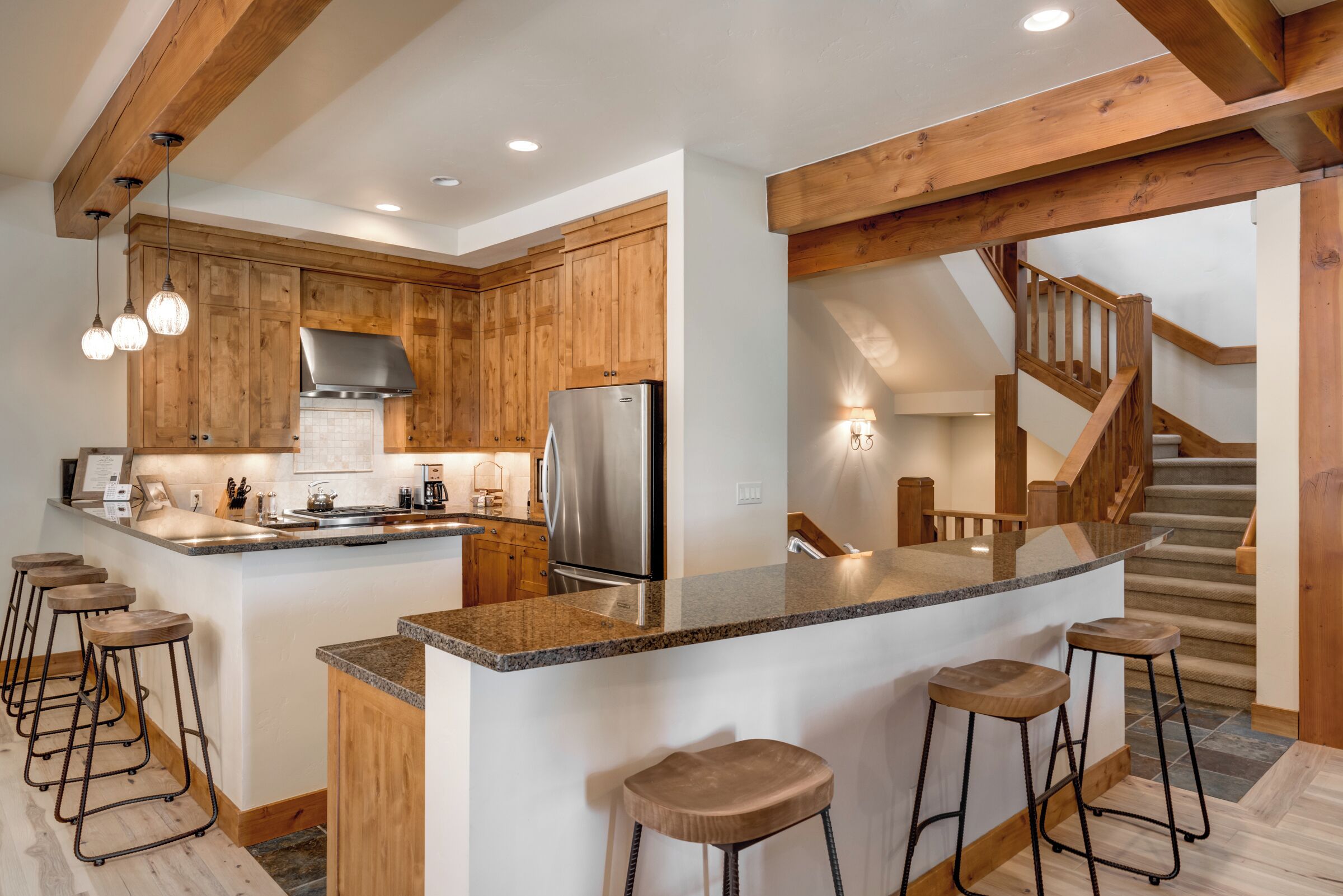 A kitchen with wooden cabinets, a stainless steel refrigerator, a granite countertop, and bar stools.