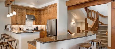 A kitchen with wooden cabinets, a stainless steel refrigerator, a granite countertop, and bar stools.