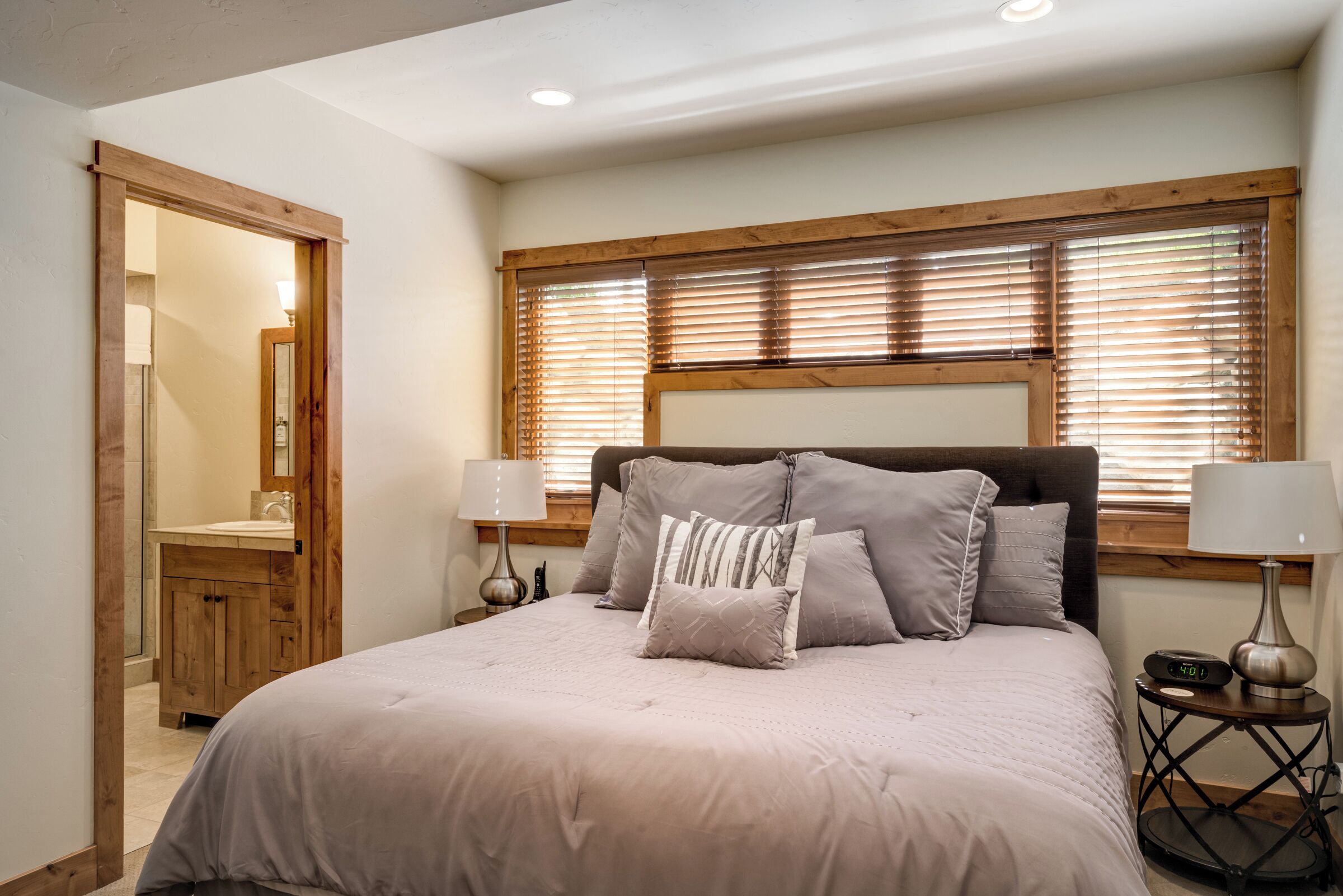 A bedroom with a bed, wooden headboard, two bedside tables with lamps, a window with blinds, and a wooden door leading to a bathroom.