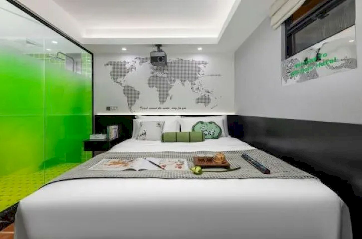 A modern hotel room with a bed, a world map on the wall, and a green glass partition.