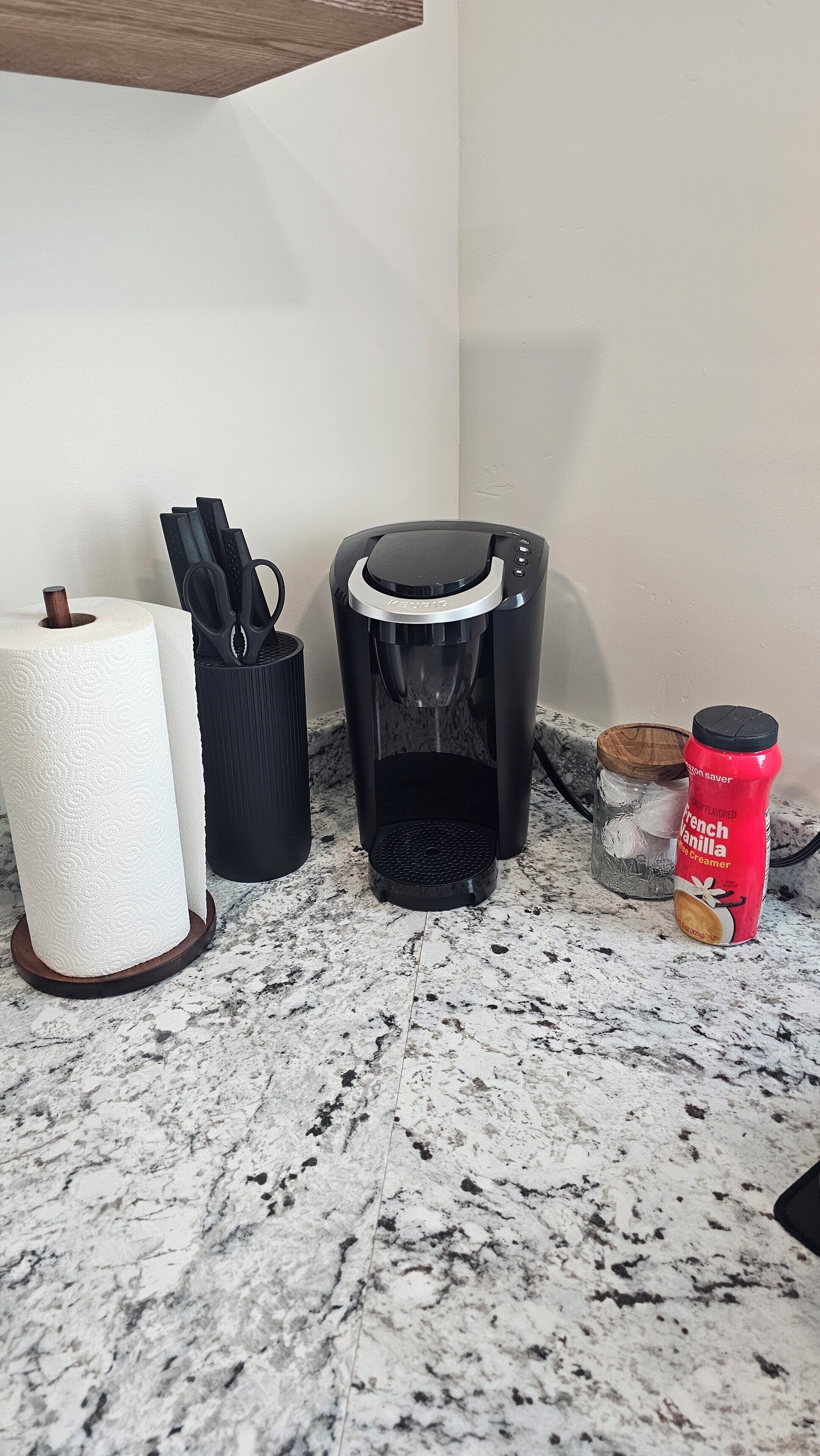A kitchen counter with a coffee maker, a roll of paper towels, a black container, a red condiment bottle, and a jar with a wooden lid.