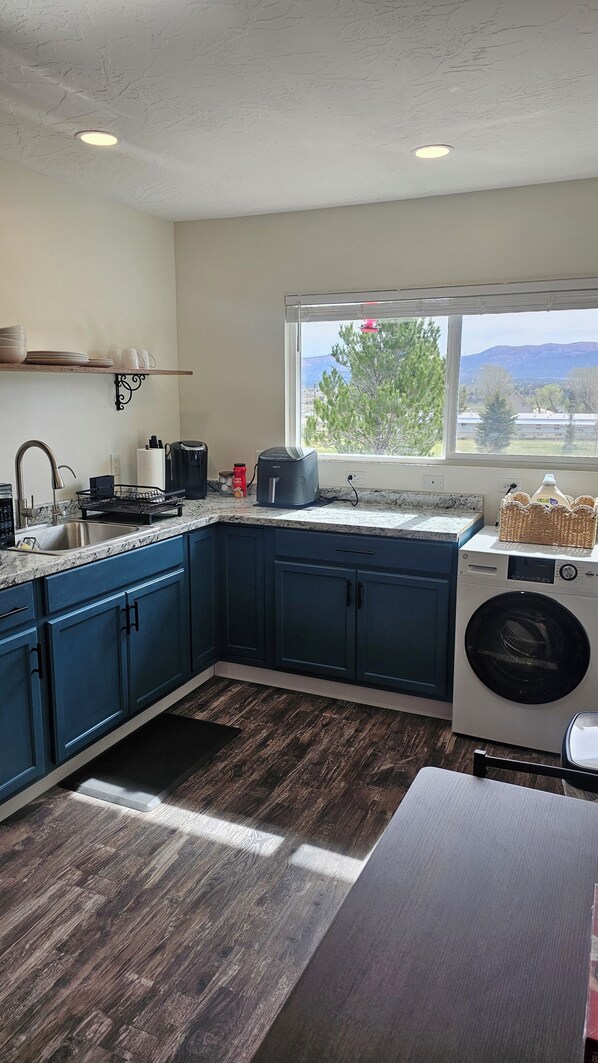 Une cuisine avec des meubles de couleur bleu foncé, un lave-linge encastré, une fenêtre donnant sur des arbres et des montagnes, et un plan de travail sur lequel se trouvent divers appareils et objets.