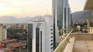 A high-rise building with a glass facade, a balcony with a yellow railing, and a view of a cityscape with mountains in the background.